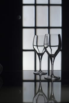 Water or wine glasses in dark room in silhouette with glass door behind defocused. Monochrome color digital photograph.