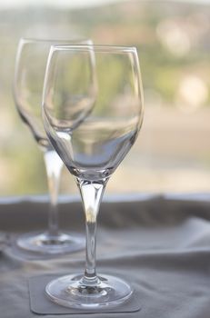 Still life photo of two wine or water glasses on coasters on cloth on tray on a table in room with window behind in soft evening pastel color light out of focus in background. 