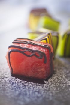 Watermelon, melon and pineapple cubes of fresh fruit cut coated in chocolate stripes dessert on dark plate with white icing sugar sprinkled. Close-up still life photo with shallow depth of focus. 