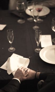 Color artistic digital rectangular vertical photo of the bride and groom holding hands on the white tablecloth of the banquet dinner table with plates and glasses in a wedding party in a luxury hotel in Spain. Shallow depth of with background out of focus. 