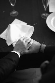 Black and white artistic digital rectangular vertical photo of the bride and groom holding hands on the white tablecloth of the banquet dinner table with plates and glasses in a wedding party in a luxury hotel in Spain. Shallow depth of with background out of focus. 