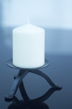 One large white candle on black metal decorative dish on plain studio blue background with reflection.
