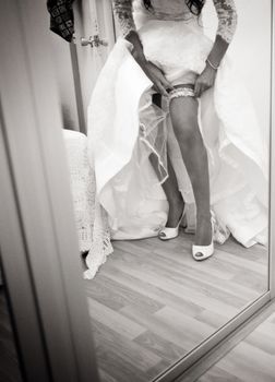 Bride on wedding day in white dress putting on garter belt shwing legs black and white photo.