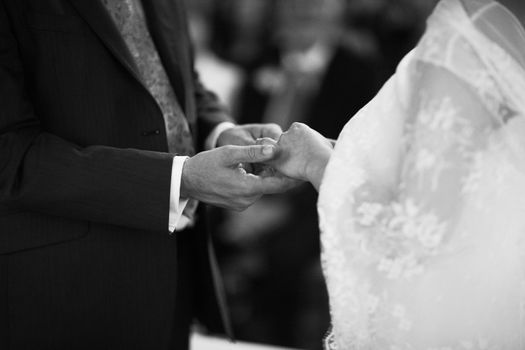 Black and white artistic digital rectangular horizontal photo of hand of bridegroom in dark long morning suit and white shirt with cufflinks in church religious wedding marriage ceremony holding hands to exchange wedding rings with the bride in white long wedding bridal dress in Barcelona Spain. Shallow depth of with background out of focus. 