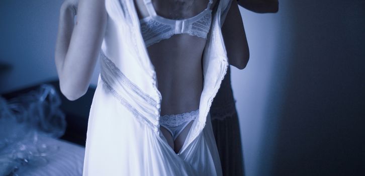 Young sensual bride in wedding dressing putting on garter belt stockings lingerie in bedroom in in blue evening light showing back and top of bottom.