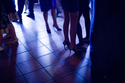 Legs of young ladies wearing high heels shoes and short cocktail dresses standing on shiny tile floor in social event wedding party in Madrid Spain. Blue evening tone color photograph. 