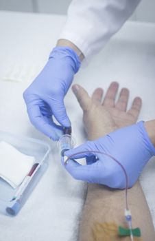 Close-up color photo of a female hospital clinic nurse wearing sterile blue gloves and senior male patient aged 65-70 giving a blood donation sample from the interior of his elbow resting arm on sterile blue white defocused background.