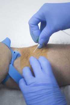 Close-up color photo of a female hospital clinic nurse wearing sterile blue gloves and senior male patient aged 65-70 giving a blood donation sample from the interior of his elbow resting arm on sterile blue white defocused background.