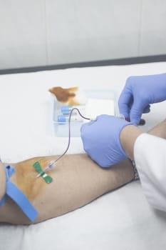 Close-up color photo of a female hospital clinic nurse wearing sterile blue gloves and senior male patient aged 65-70 giving a blood donation sample from the interior of his elbow resting arm on sterile blue white defocused background.