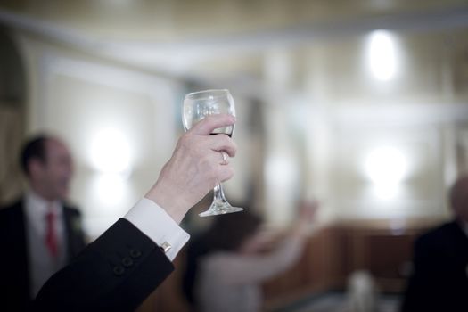 Color artistic digital rectangular horizontal photo of hand of man in dark suit and white shirt in wedding reception banquet marriage party holding  glass of red wine glass. Shallow depth of with background out of focus. 