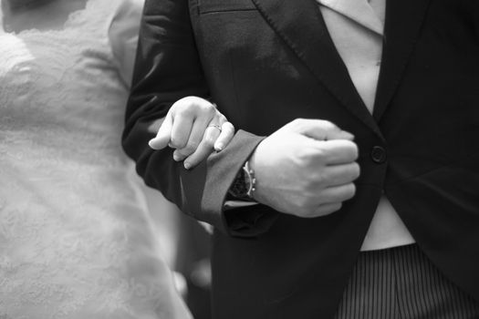 Black and white artistic digital photo of bridegroom in dark suit and white shirt in wedding marriage holding hands with the bride in white long wedding bridal dress. Shallow depth of with background out of focus. 