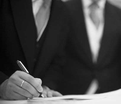 Black and white artistic digital rectangular horizontal photo of hand of bridegroom with wedding ring holding pen in dark suit and white shirt in wedding signing marriage register in Barcelona Spain. Shallow depth of with background out of focus. 