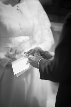 Black and white artistic digital photo of bridegroom in dark suit and white shirt in church religious wedding marriage ceremony holding hands with the bride in white long wedding bridal dress. Shallow depth of with background out of focus. 