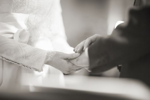 Black and white artistic digital photo of bridegroom in dark suit and white shirt in church religious wedding marriage ceremony holding hands with the bride in white long wedding bridal dress. Shallow depth of with background out of focus. 