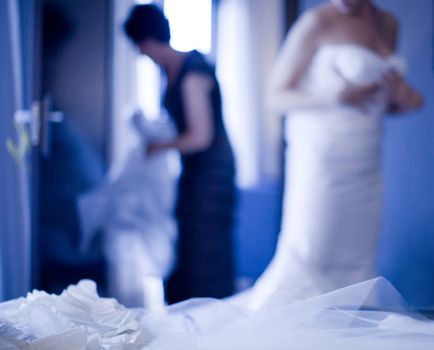 Bride in wedding dressing putting on garter belt stockings lingerie in bedroom of hotel with help of mother in Madrid Spain in blue evening light defocused with focus on white garter belt on bed in foreground.