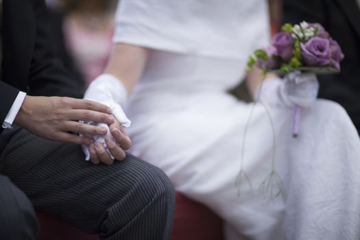 Color artistic digital photo of bridegroom in dark suit and white shirt with cufflinks in church religious wedding marriage ceremony holding hands with the bride in white wedding bridal dress. Shallow depth of with background out of focus. 