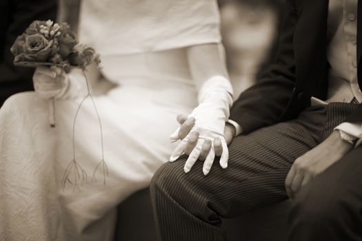 Black and white artistic digital photo of bridegroom in dark suit and white shirt in church religious wedding marriage ceremony holding hands with the bride in white long wedding bridal dress. Shallow depth of with background out of focus. 