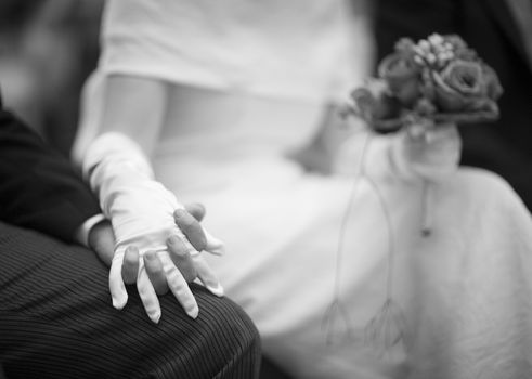 Black and white artistic digital photo of bridegroom in dark suit and white shirt in church religious wedding marriage ceremony holding hands with the bride in white long wedding bridal dress. Shallow depth of with background out of focus. 