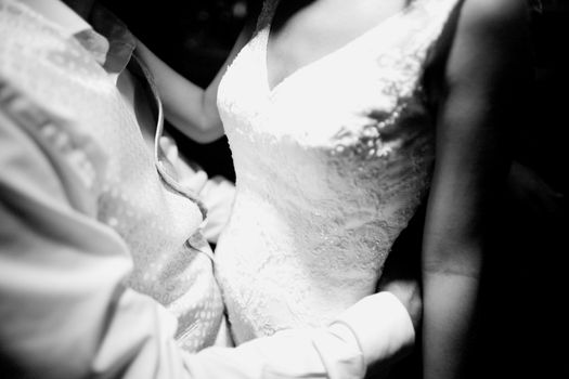 Black and white artistic digital photo of bridegroom in dark suit in wedding marriage event holding hands with the bride in white long wedding bridal dress. Shallow depth of with background out of focus. 