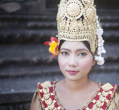 Siem Reap, Cambodia ��� November 27, 2013: Young Cambodian lady dancer in the street wearing traditional Cambodian red and gold dress costume, makeup, ceremonial gold color metal headwear and Cambodian flowers outside Angkor Wat ancient ruined temples looking at the camera before starting traditional dance routine done to earn money from international tourists. Color portrait photo in of a female dancer in the street in Siem Reap Cambodia by Angkor Wat temple ruins in traditional Cambodian dress and headwear with face makeup looking at the camera.

Color portrait photo in of a female dancer in the street in Siem Reap Cambodia by Angkor Wat temple ruins in traditional Cambodian dress and headwear with face makeup looking at the camera. 