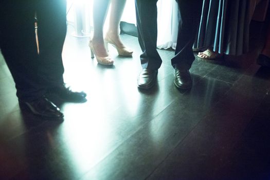Legs of young lady wearing high heels shoes and men in suits standing on shiny tile floor in social event wedding marriage party in Madrid Spain. Green night tone color photograph. 