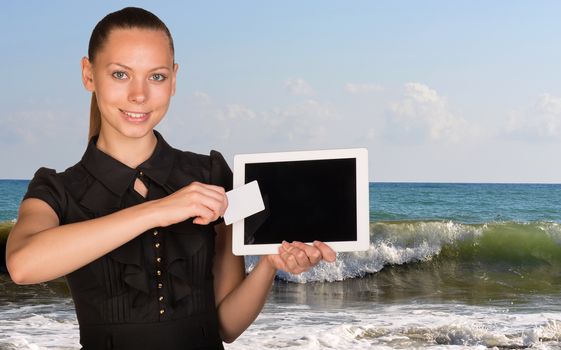 Beautiful businesswoman holding blank tablet PC and blank business card in front of PC screen. Seascape whith tidal waves as backdrop