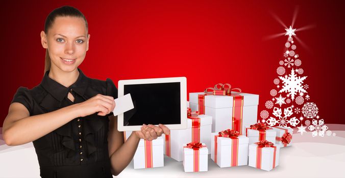 Beautiful businesswoman holding blank tablet PC and blank business card in front of PC screen. New Year tree composed of snowflakes and gifts clustered under the tree as backdrop