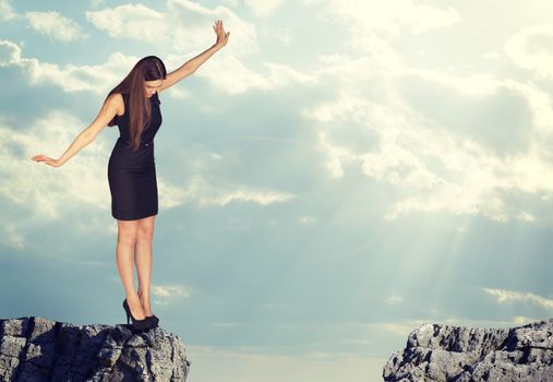 Businesswoman with her arms outstretched as for balance standing on the edge of rock gap and looking down into it. Sky and clouds as backdrop