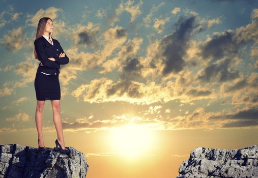 Businesswoman standing on the edge of rock gap, with her arms crossed on her breast as if uncertain. Sunrise sky as backdrop