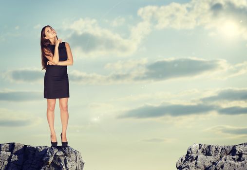 Businesswoman standing on the edge of rock gap in thoughtful pose. Sky and clouds as backdrop