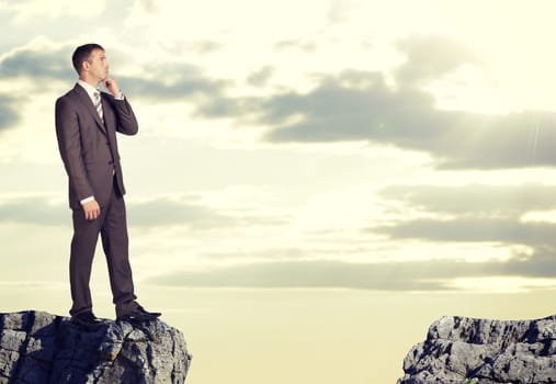 Businessman standing on the edge of rock gap in thoughtful pose. Sky and clouds as backdrop