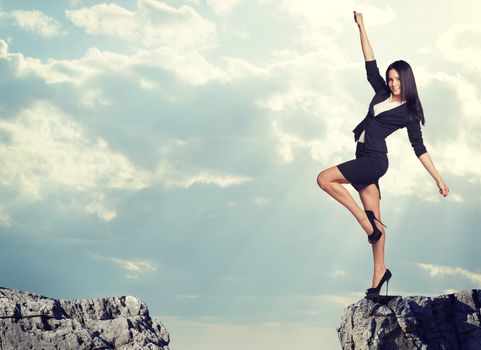 Businesswoman standing on the edge of rock gap in cheerful pose as if going to leap across it. Sky and clouds as backdrop