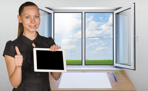 Beautiful businesswoman holding blank tablet PC and giving thumb up. Open window with green meadow and sky as backdrop