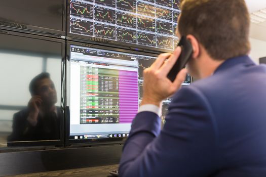Businessman with cell phone trading stocks. Stock analyst looking at graphs, indexes and numbers on multiple computer screens. Stock trader evaluating economic data.                  