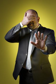 Stressed businessman in dark suit holding up hand in front of his eyes and parries with the other hand, against yellow background