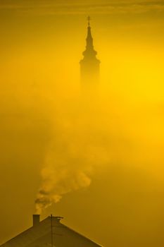 Golden sunrise church tower in fog and chimney smoke in town of Krizevci, Prigorje region, Croatia