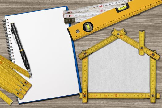 House project concept. Wooden meter ruler in the shape of house with pencil, empty notebook, two meter tools and spirit level on a wooden desk