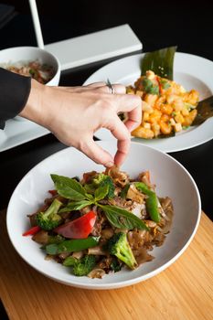 Food stylist grooms a dish by adding sweet basil garnish to the traditional thai dish Pad Kee Mao drunken noodle.