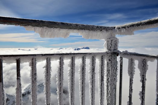 Horizontal landscape of a frost and snow covered balcony in the foreground and a thick layer of clouds and some mountaintops behind it against blue sky.