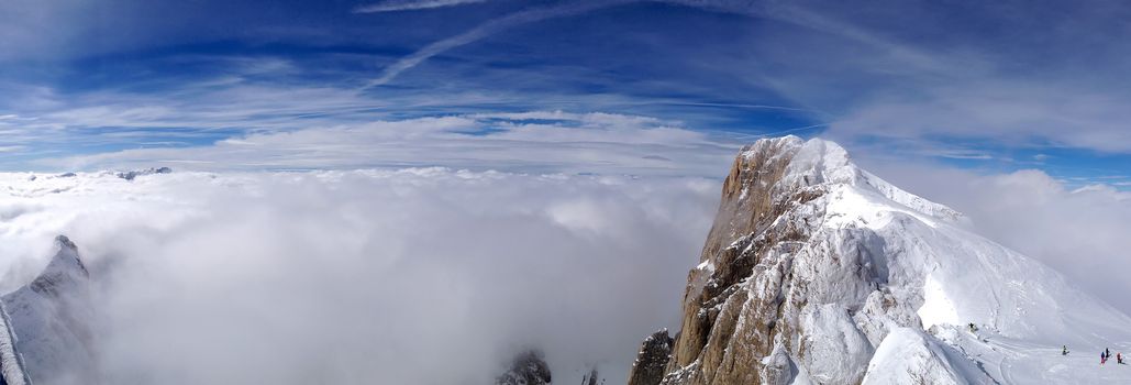 Horizontal panorama of a snow covered alpine mountain surrounded by a thick layer of clouds, beneath a vibrant blue sky.