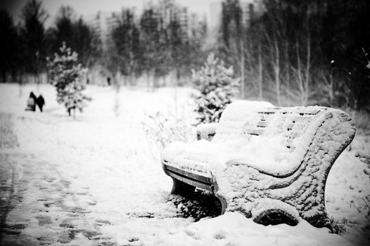 Horizontal black and white landscape of a snow covered bench in the winter park.