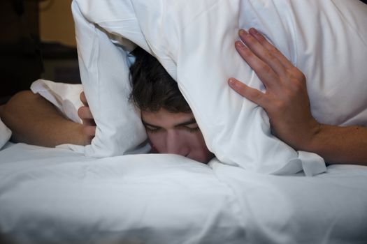 Handsome young male insomniac covering his head and ears trying to block out the sound with a pillow as he is kept awake by loud noises around him