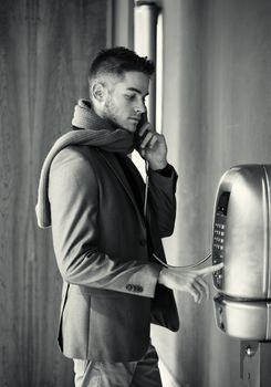 Handsome stylish young man in a tailored jacket and scarf standing using a pay phone waiting for a connection, profile view in greyscale