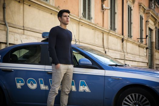 Handsome trendy young Italian policeman standing alongside a patrol car parked outside an old urban building watching something off frame to the right