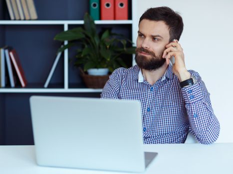 Young bearded man working on a laptop in the office and talking on cell phone