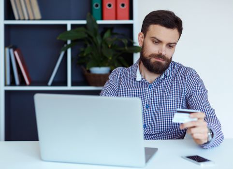 Young bearded man working on a laptop in the office - pays by credit card online shopping