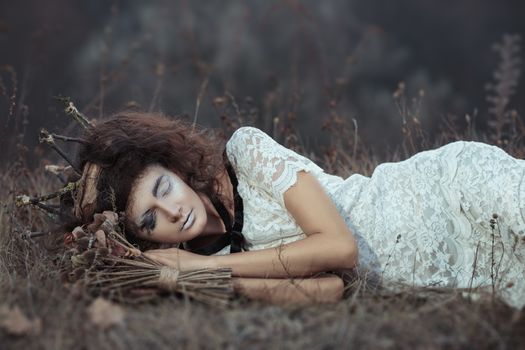 Young girl in the image of a bride in a white lace dress, a crown of twigs with a bouquet of dried flowers lying on the ground