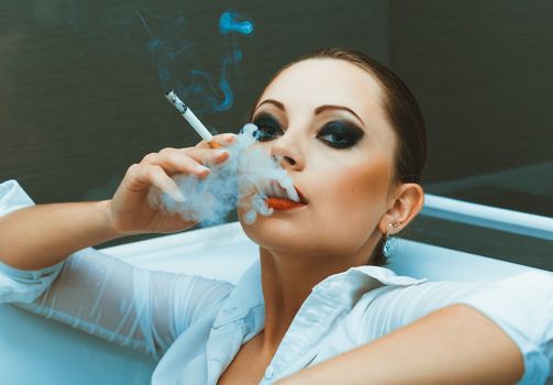 Young attractive girl in a white shirt with bright makeup lying in the bathroom and smokes a cigarette