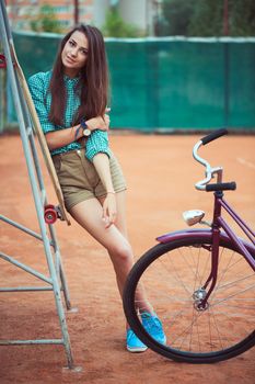 Beautiful young girl with longboard and bicycle standing on the tennis court, outdoor