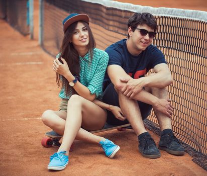 Young beautiful couple sitting on a skateboard on the tennis court